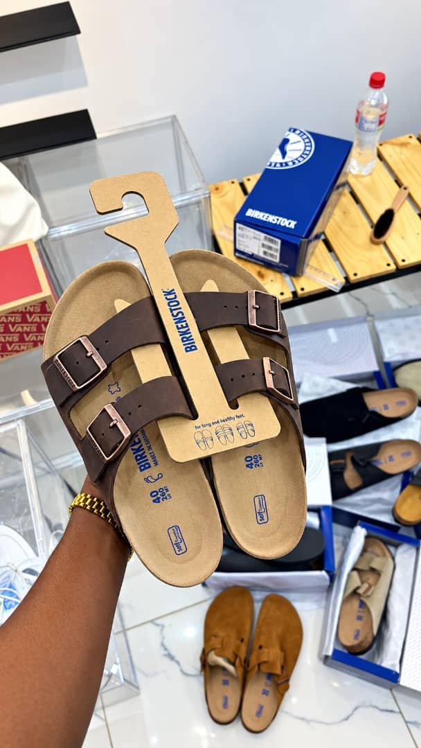 root A pair of brown Birkenstock sandals with two adjustable buckles, presented on a cardboard holder, showcasing Modern Luxury and Urban Edge.