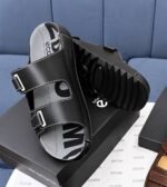 A pair of black leather slides with two adjustable straps and a thick, ridged sole, resting on a shoe box.