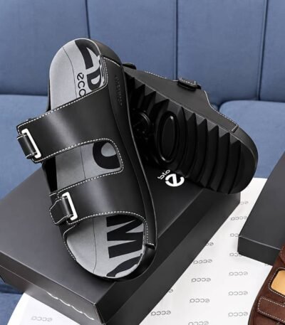 A pair of black leather slides with two adjustable straps and a thick, ridged sole, resting on a shoe box.
