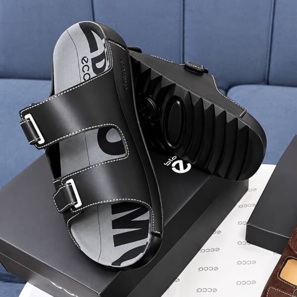 A pair of black leather slides with two adjustable straps and a thick, ridged sole, resting on a shoe box.