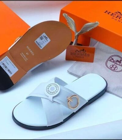 A pair of white leather slides with a gold-tone buckle, displayed with an orange box and dust bag, showcasing Modern Luxury and Timeless Quality.