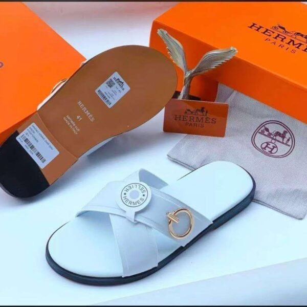 A pair of white leather slides with a gold-tone buckle, displayed with an orange box and dust bag, showcasing Modern Luxury and Timeless Quality.