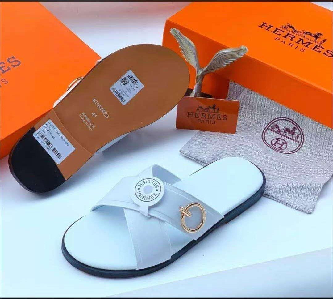 root A pair of white leather slides with a gold-tone buckle, displayed with an orange box and dust bag, showcasing Modern Luxury and Timeless Quality.