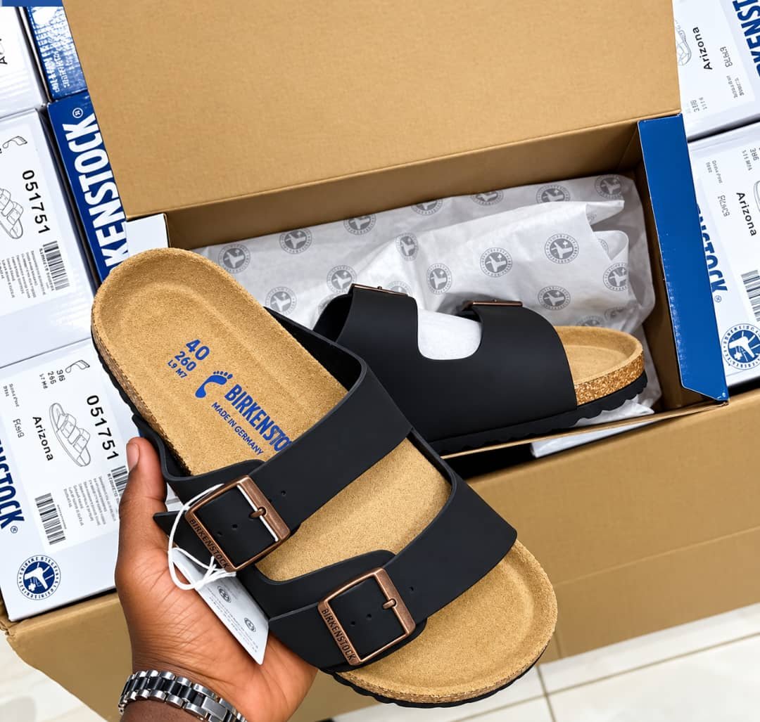 root Birkenstock Arizona Slide in black, featuring a contoured cork footbed and two adjustable buckle straps, presented in its packaging.