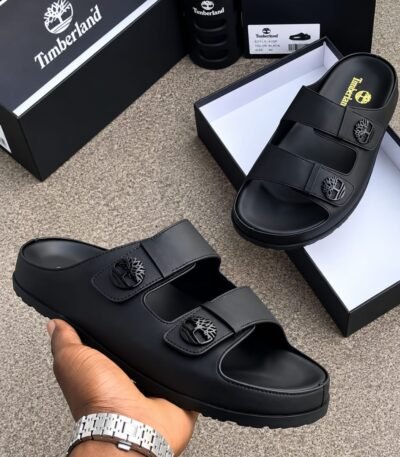 Timberland black slides with two wide straps and a tree logo on each strap, displayed on a gray surface with boxes and a hand holding one slide.