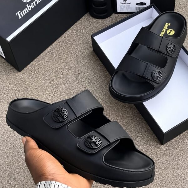 Timberland black slides with two wide straps and a tree logo on each strap, displayed on a gray surface with boxes and a hand holding one slide.