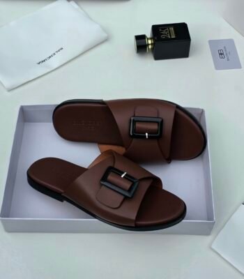 A pair of luxurious brown leather sandals with black buckles, nestled in a white box alongside a white cloth and a sleek black perfume bottle, exuding sophistication and refinement.