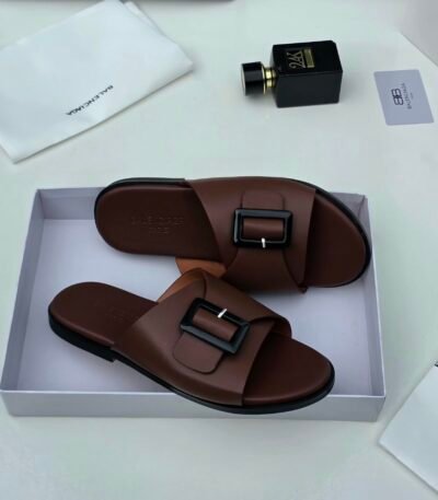 A pair of luxurious brown leather sandals with black buckles, nestled in a white box alongside a white cloth and a sleek black perfume bottle, exuding sophistication and refinement.