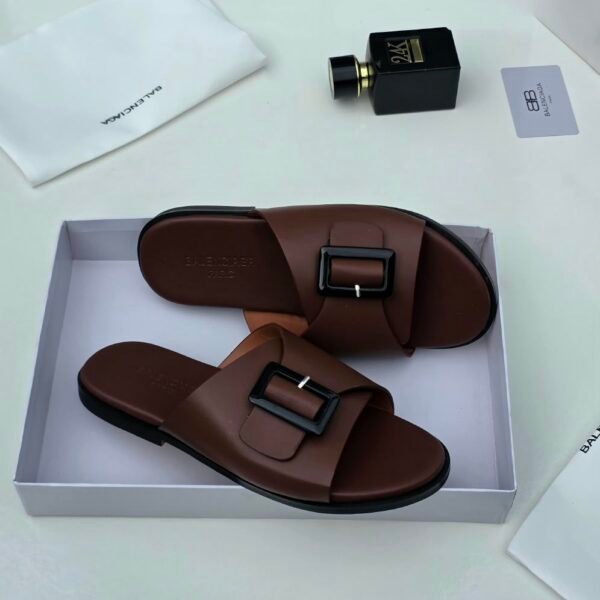 A pair of luxurious brown leather sandals with black buckles, nestled in a white box alongside a white cloth and a sleek black perfume bottle, exuding sophistication and refinement.