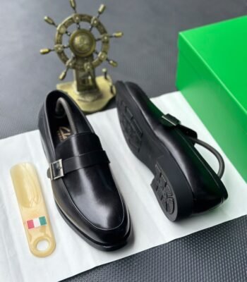 A pair of sleek black leather loafers with a stylish buckle, presented on a crisp white surface alongside a green box and a golden ship's wheel, evoking a sense of luxury and refinement.