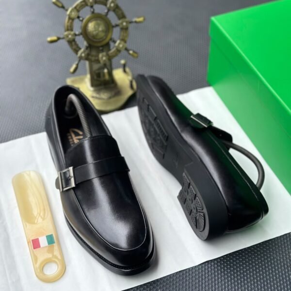 A pair of sleek black leather loafers with a stylish buckle, presented on a crisp white surface alongside a green box and a golden ship's wheel, evoking a sense of luxury and refinement.