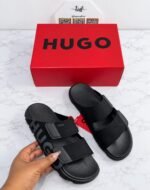 A pair of tan-colored HUGO slides with two wide straps and a chunky sole, set against a clean white background.