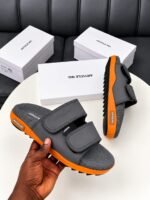 A pair of black slides with orange soles, presented on a white table with white boxes in the background, embodying Modern Luxury and Urban Edge.