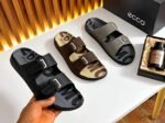 A pair of black leather slides with two adjustable straps and a thick, ridged sole, resting on a shoe box.