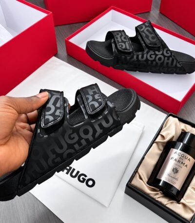 A pair of black HUGO sandals with a bold design and premium materials, presented in a luxurious setting with red boxes and high-end fragrance.