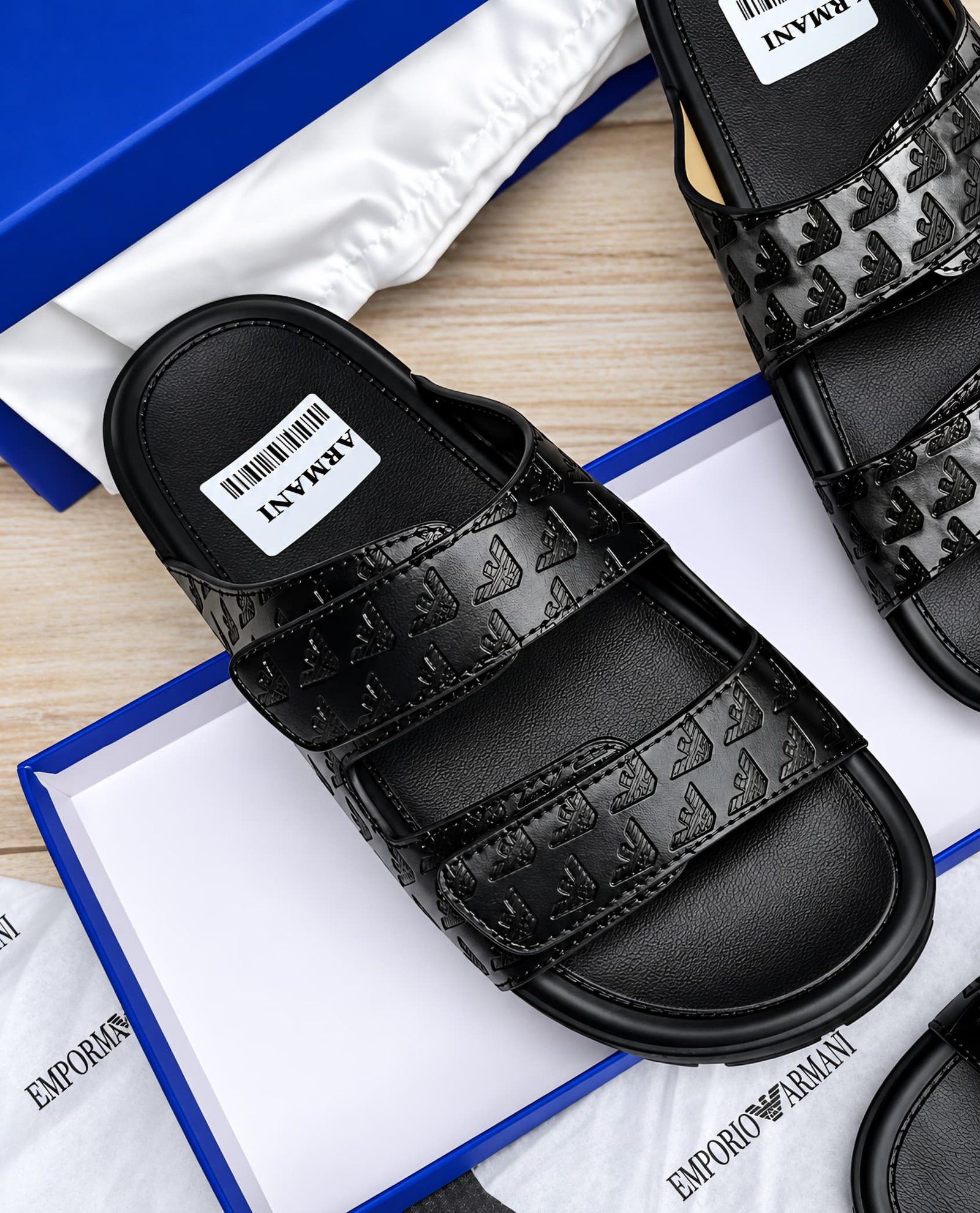 root A pair of black leather slides with debossed logo design, presented in a blue box with white packaging, showcasing Modern Luxury and Urban Edge style.