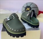 A pair of olive green clog shoes with silver studs and a metallic buckle, placed on a cream-colored shoe box with a basketball in the background.