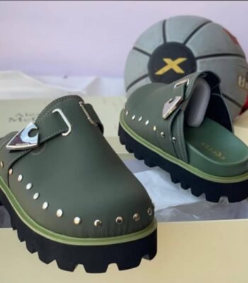 A pair of olive green clog shoes with silver studs and a metallic buckle, placed on a cream-colored shoe box with a basketball in the background.