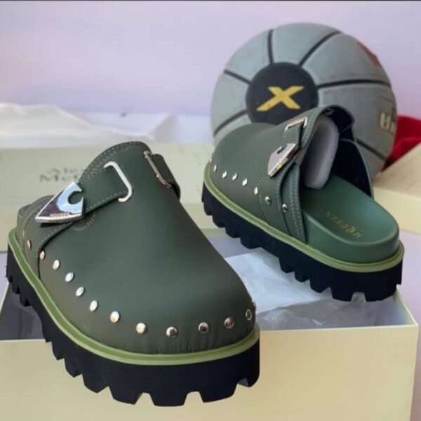 A pair of olive green clog shoes with silver studs and a metallic buckle, placed on a cream-colored shoe box with a basketball in the background.
