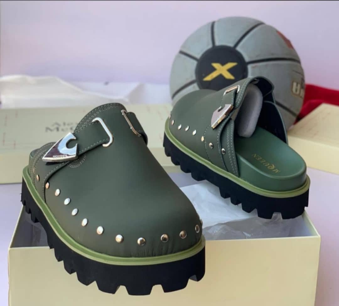 root A pair of olive green clog shoes with silver studs and a metallic buckle, placed on a cream-colored shoe box with a basketball in the background.